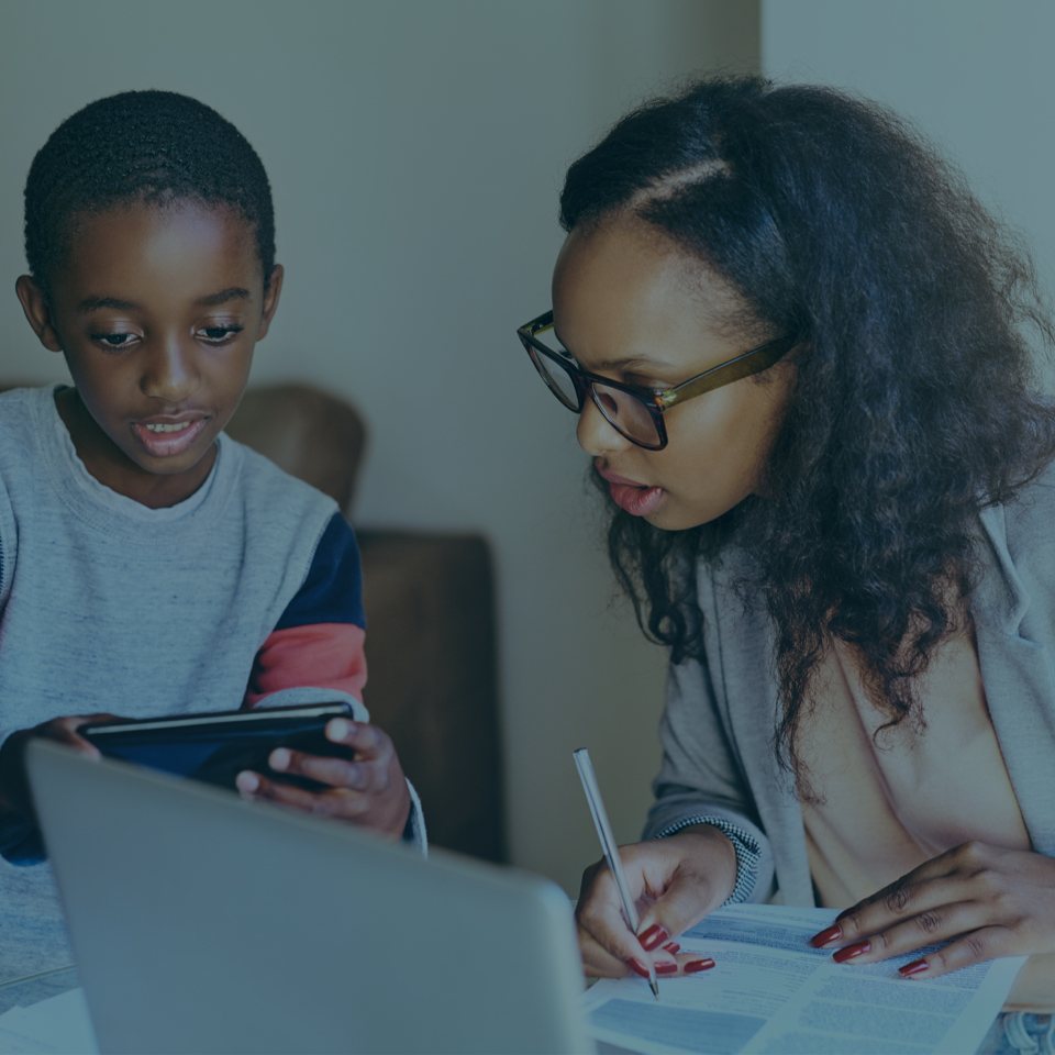 A woman wearing glasses and a gray blazer is sitting at a table with a boy. The boy is holding a tablet, and the woman is writing on a piece of paper next to a laptop. They appear to be working together in a home or office setting.