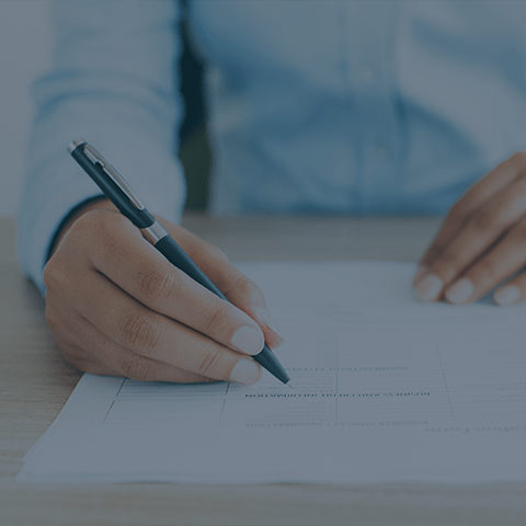 Close-up of a person in a light blue shirt, holding a pen and writing on a form or document on a wooden desk. The focus is on the hand and pen, with the rest of the person slightly out of focus in the background.