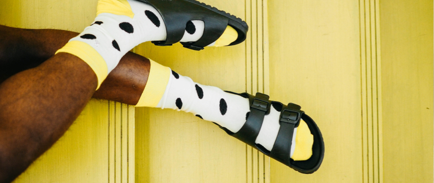 A person wearing bright yellow socks with black polka dots and black sandals rests their legs crossed against a yellow background. The image captures the contrast between the vibrant socks and the dark sandals.