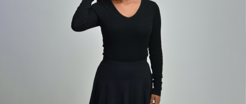 A person with shoulder-length dark hair is posing against a plain gray backdrop. They are wearing a black long-sleeve shirt and a black skirt. The person has their right hand raised to their head, touching their hair.