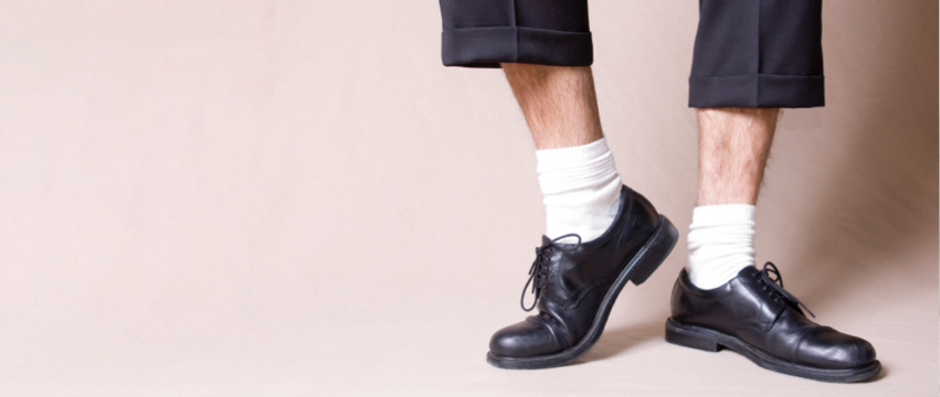 A close-up shot of a person wearing black dress shoes with white socks and dark rolled-up pants. The person’s feet are angled slightly, and the background is a plain light beige.