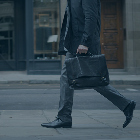 A person dressed in a business suit is walking on a city sidewalk while carrying a black leather briefcase. The background shows storefronts and an entrance to a building. The overall tone of the image is monochromatic with blue hues.