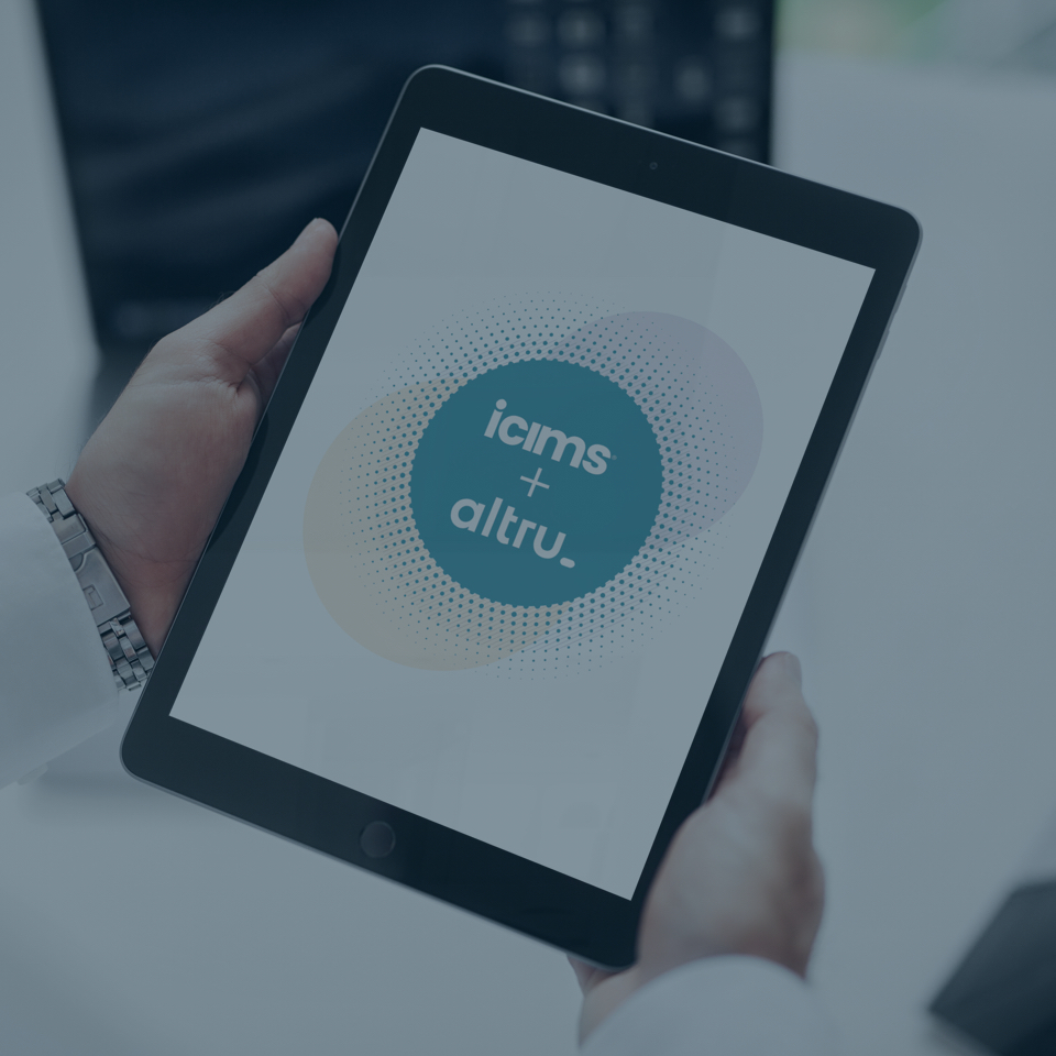 A close-up of hands holding a black tablet vertically. On the screen, a graphic displays the text "icims + altru" within overlapping, multicolored circles. The background shows a blurred office setting with a desk and open laptop.