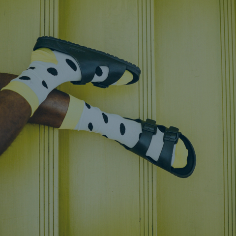 A person's legs wearing black sandals with straps and yellow socks featuring large black polka dots. The background is a light green wall with vertical grooves.