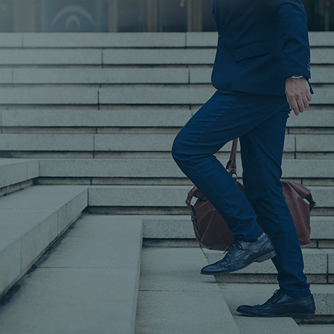 A person dressed in business attire is walking up concrete stairs. Their lower body is visible, showing blue pants, a dark blazer, and polished shoes. They are holding a brown leather bag in their left hand.