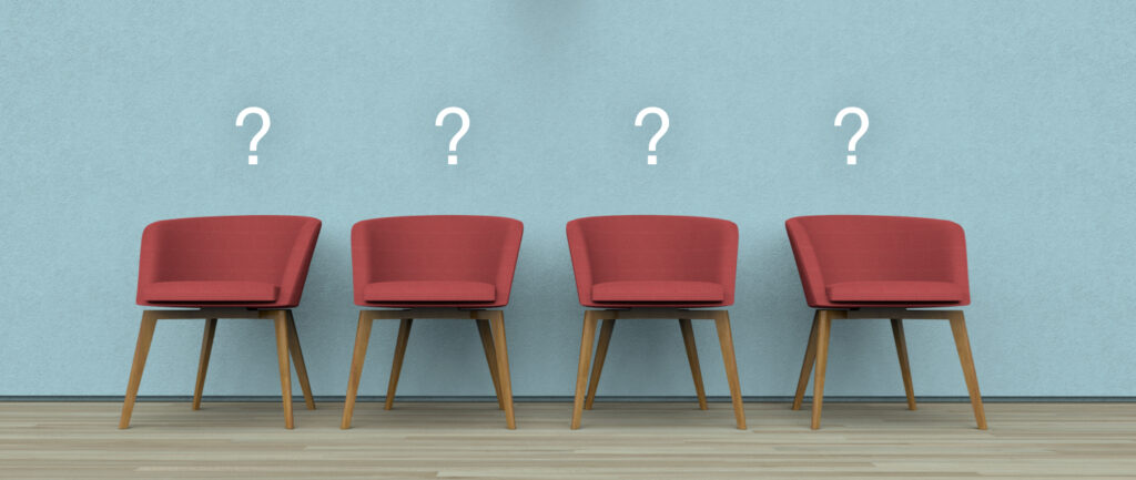 Four red chairs with wooden legs are lined up against a pale blue wall. Each chair has a white question mark above it, suggesting uncertainty or an element of questioning, possibly related to choices or decision-making. The floor is made of light wood.