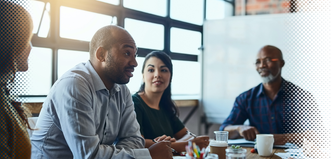 A diverse group of four people are sitting at a table in a modern office having a discussion. One person is speaking, while the others listen attentively. There is a large window in the background and a whiteboard to the side.