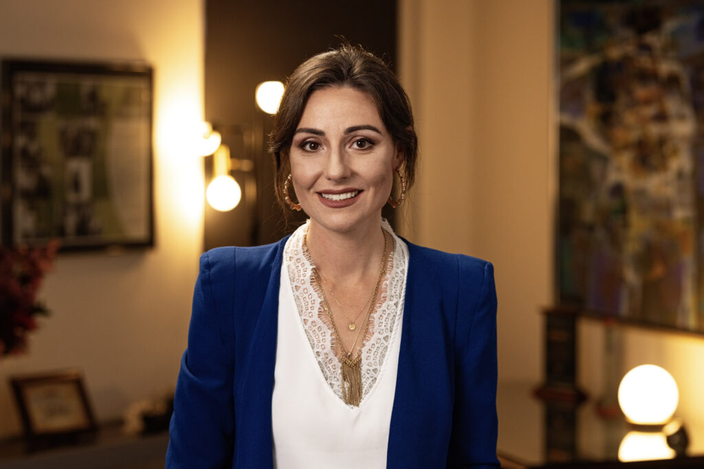 A woman with shoulder-length dark hair smiles while standing indoors. She is wearing a white blouse with a lace neckline and a blue blazer. The background is softly lit, featuring framed pictures on the walls and some illuminated decorative lights.