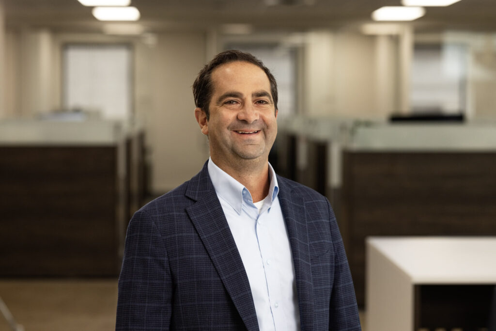 A man with short dark hair and a beard is standing in an office environment. He is wearing a dark blue blazer over a light blue shirt and is smiling at the camera. The office has cubicles with glass dividers and is brightly lit.