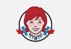 Illustration of a girl with red hair styled in pigtails, secured with light blue bows. She has a face full of freckles and a wide smile. She is wearing a white collared shirt with blue stripes. The image is outlined in a thin black circle.