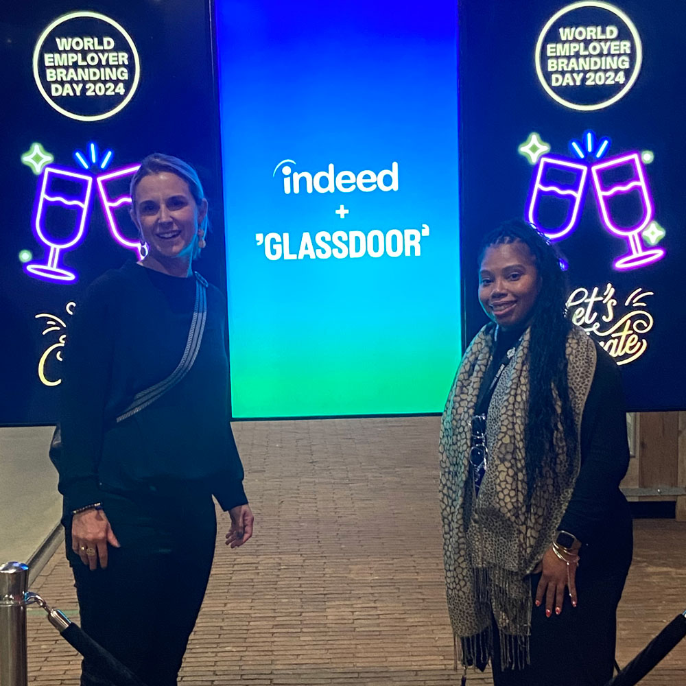Two people smile in front of a colorful backdrop with the logos of Indeed and "Glassdoor." Neon signs reading "World Employer Branding Day 2024" and "Let's Celebrate" are visible on the sides.