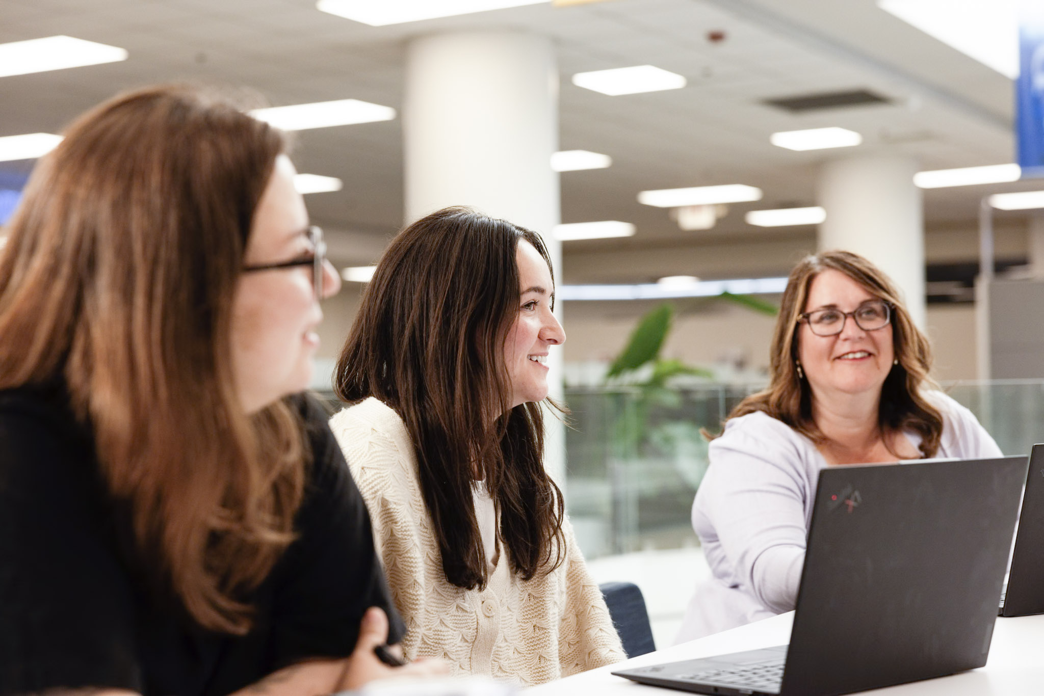 Three women are sitting at a table indoors, smiling and talking. Two of them have laptops open in front of them, and the background shows a bright, modern office space with large pillars.