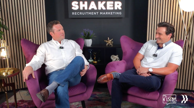 Two men sit in purple armchairs, engaged in conversation. They wear microphones and casual business attire. Behind them is a sign reading "SHAKER Recruitment Marketing" on a dark wall with decorative items and plants.