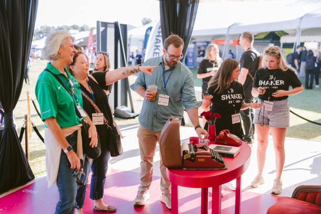 A group of people interact around a round red table with vintage phones at an outdoor event. Some wear name badges while others wear matching black "SHAKER" shirts. Tents and other attendees are visible in the background.