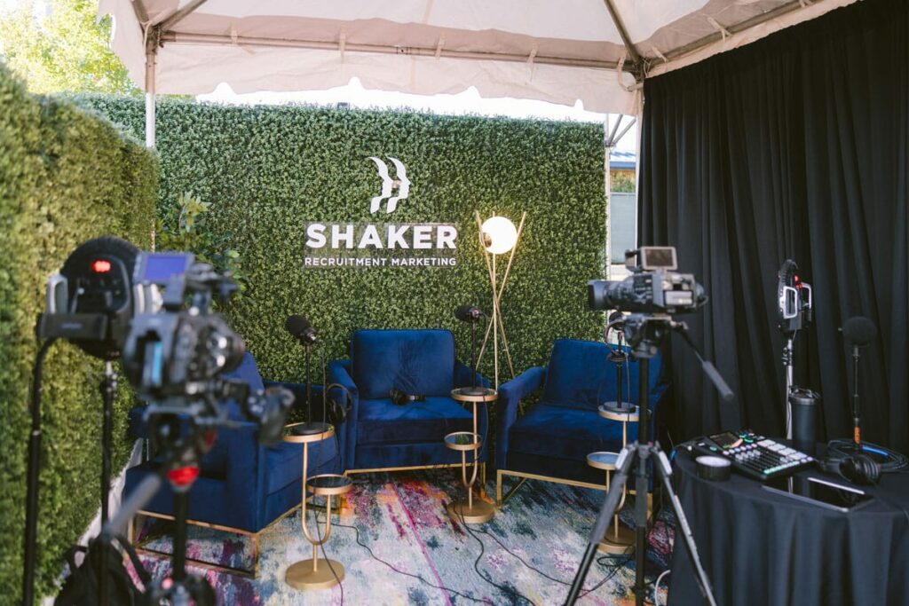 A podcast or interview setup with blue chairs, microphones, cameras, and lighting, under a tent. The backdrop features green foliage with the "SHAKER Recruitment Marketing" logo.