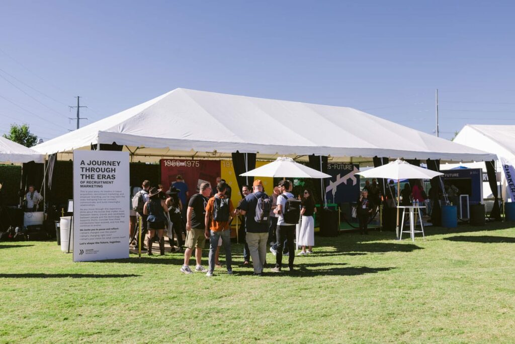 A group of people stand and talk outside a large white event tent on a sunny day. The tent features displays inside and a sign reading “A Journey Through the Eras.” Green grass and clear blue sky are visible.