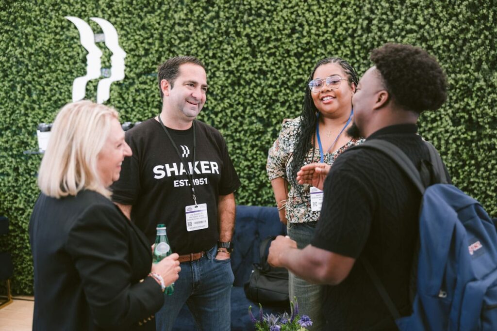 Four people stand in a circle having a friendly conversation at an outdoor event, with a green leafy wall in the background. One person holds a drink; others are wearing badges and are smiling and laughing.