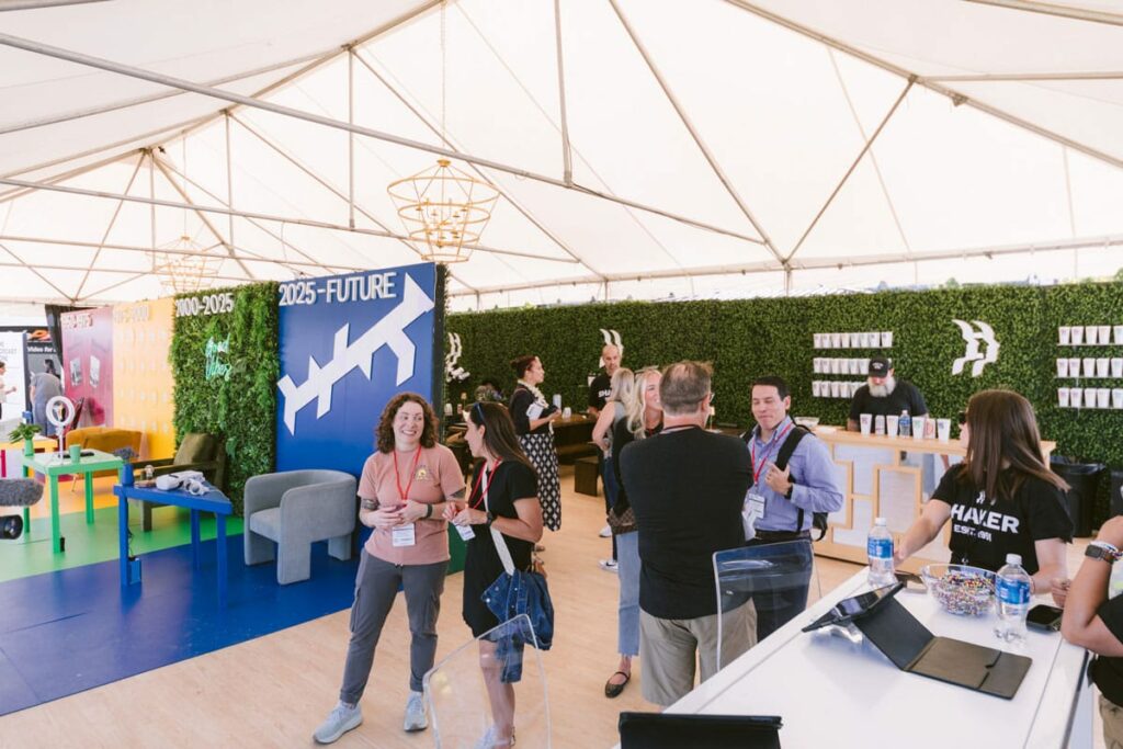 People converse and network inside a large event tent with lounge areas, colorful displays, and a green wall featuring the text "2025=FUTURE." Lanyards and event badges are visible on several attendees.