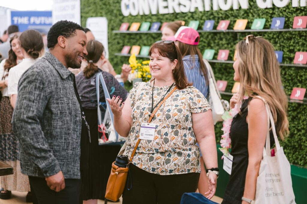 Three people stand and smile while having a conversation at an event. Other attendees and a display with colorful cards labeled “CONVERSATION GUIDES” are visible in the background.