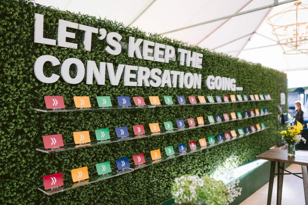 A green hedge wall displays the large white text “LET’S KEEP THE CONVERSATION GOING…” above several rows of colorful cards placed on clear shelves, inside a bright event tent.
