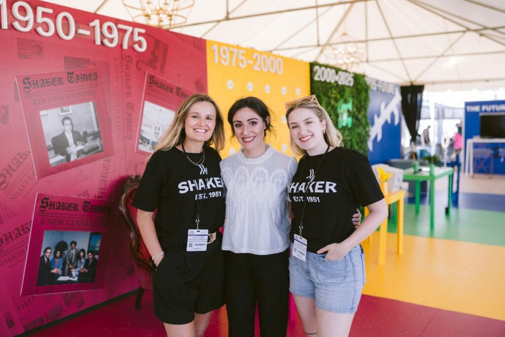 Three smiling women stand together inside a colorful, tented exhibit displaying Shaker history from 1950 to 2015. Two wear black "SHAKER EST. 1951" shirts, and all wear event badges.