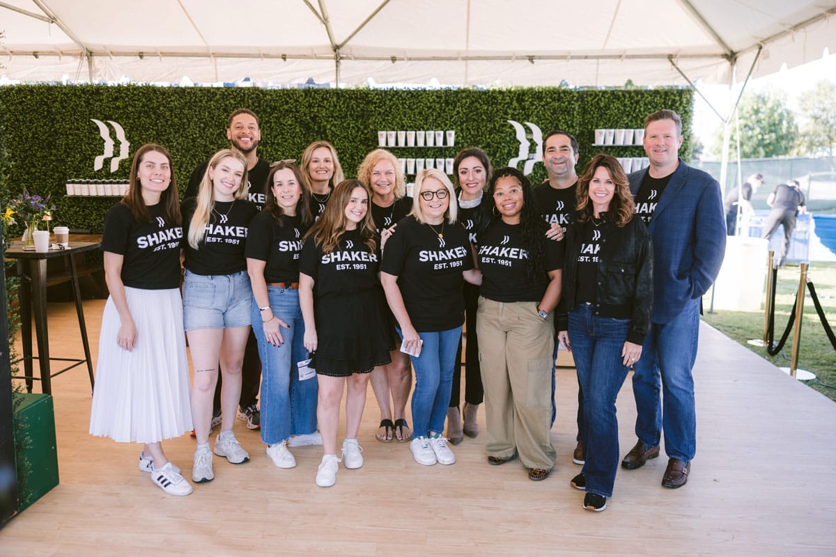 A group of thirteen people stand together smiling under a tent. Most wear matching black “SHAKER” shirts, while a few are in business or casual attire. A green hedge wall with white logos is in the background.