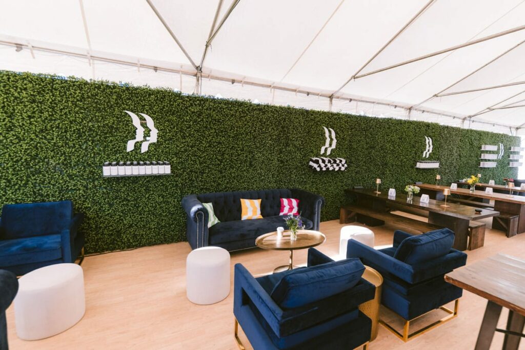 Modern lounge area with blue velvet sofas, colorful pillows, white ottomans, and a gold coffee table, set against a green hedge wall with minimalist white shelves under a large white tent.