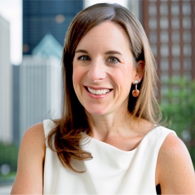 A woman with long brown hair, wearing a sleeveless white top and drop earrings, smiles at the camera. She stands outdoors with blurred city buildings in the background.