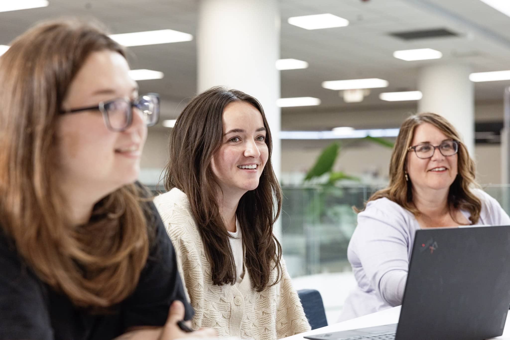 Three women sit at a table in a bright, modern office space, engaged in conversation. One woman is using a laptop, and all three appear to be smiling and paying attention.