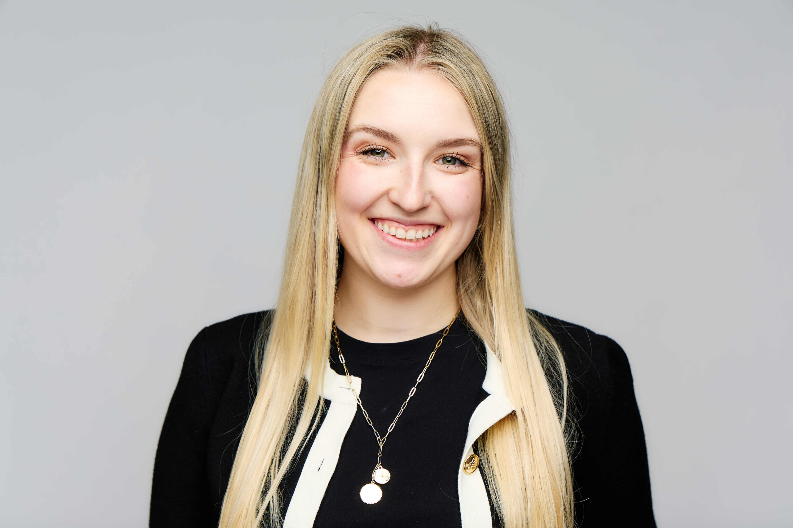 A young woman with long blonde hair smiles at the camera. She is wearing a black top, a black and white jacket, and layered gold necklaces. The background is plain light gray.