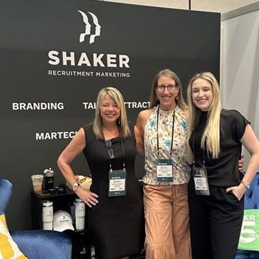 Three women stand smiling together in front of a booth display for "Shaker Recruitment Marketing," with words like "Branding," "Talent Attract," and "Martech" visible on the backdrop.