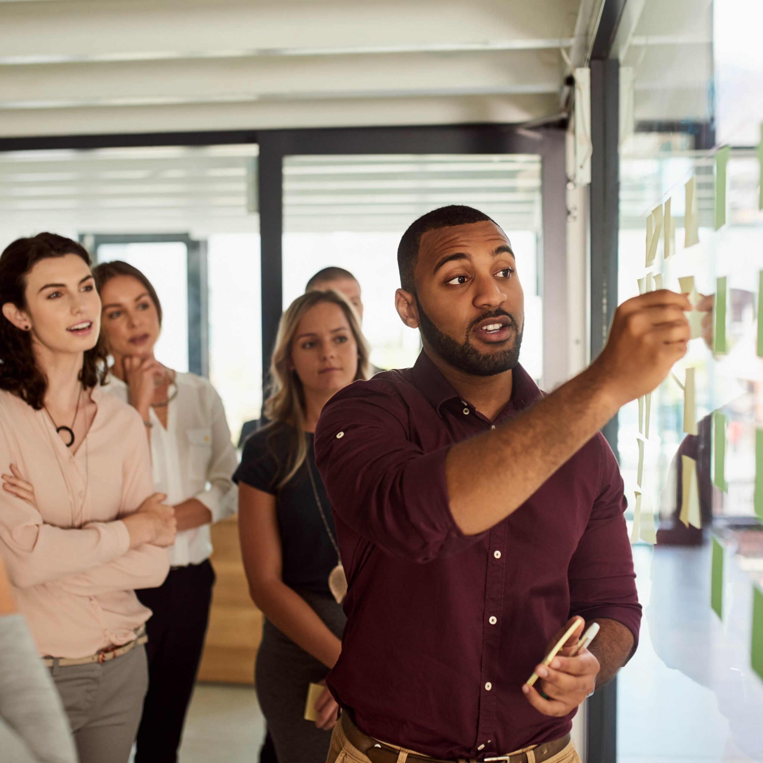 A man in a maroon shirt places sticky notes on a glass wall while four colleagues watch attentively in a bright office setting.