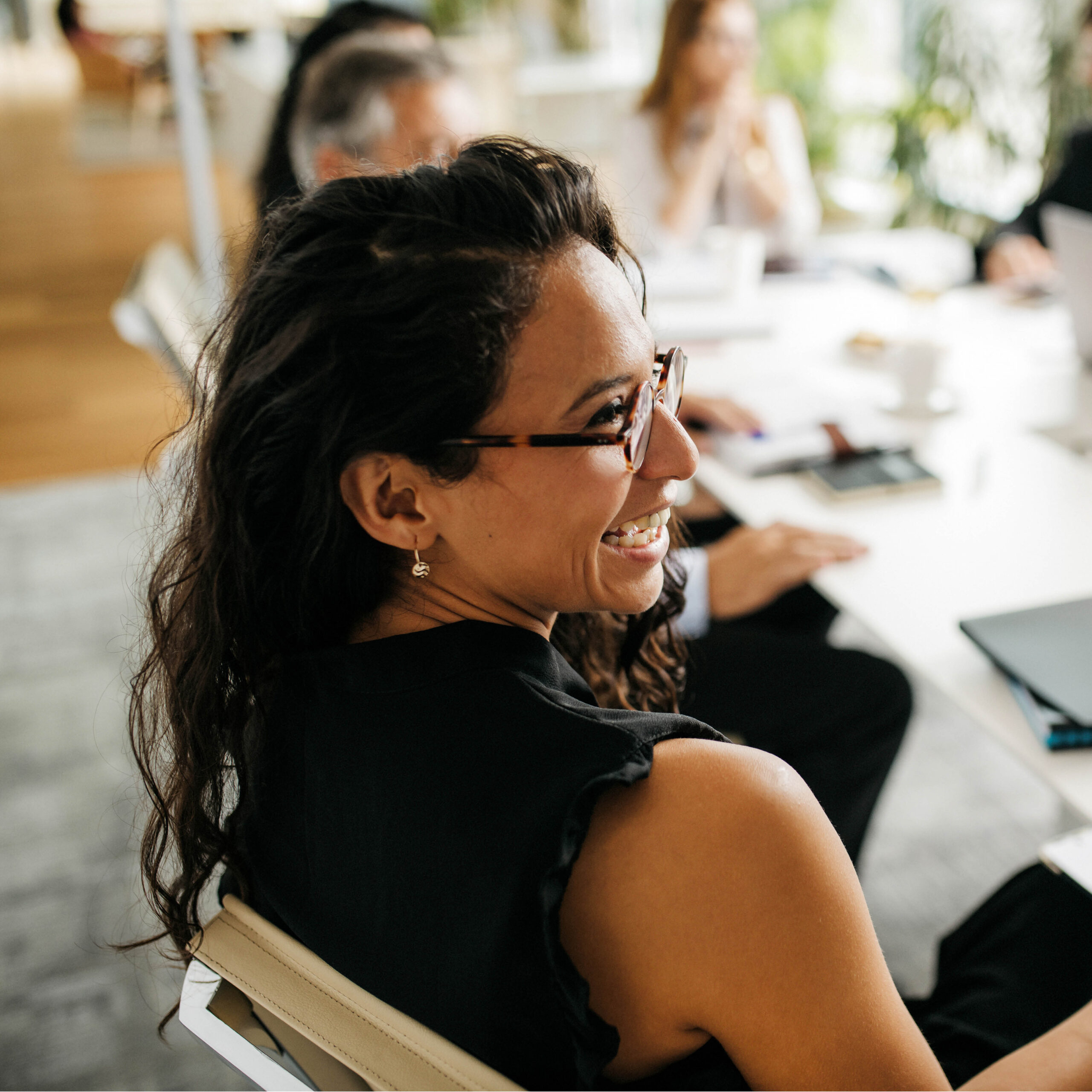 A woman with long curly hair and glasses smiles while sitting at a meeting table with others. She is wearing a sleeveless black top, and the background shows blurred colleagues and office items.