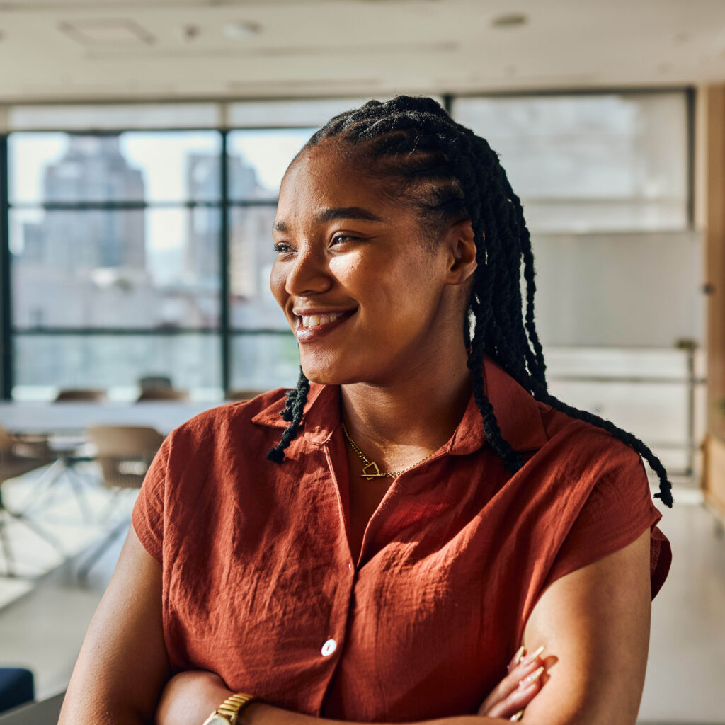 A woman with braided hair wearing a rust-colored short-sleeve shirt stands smiling with arms crossed in a bright, modern office with large windows in the background.