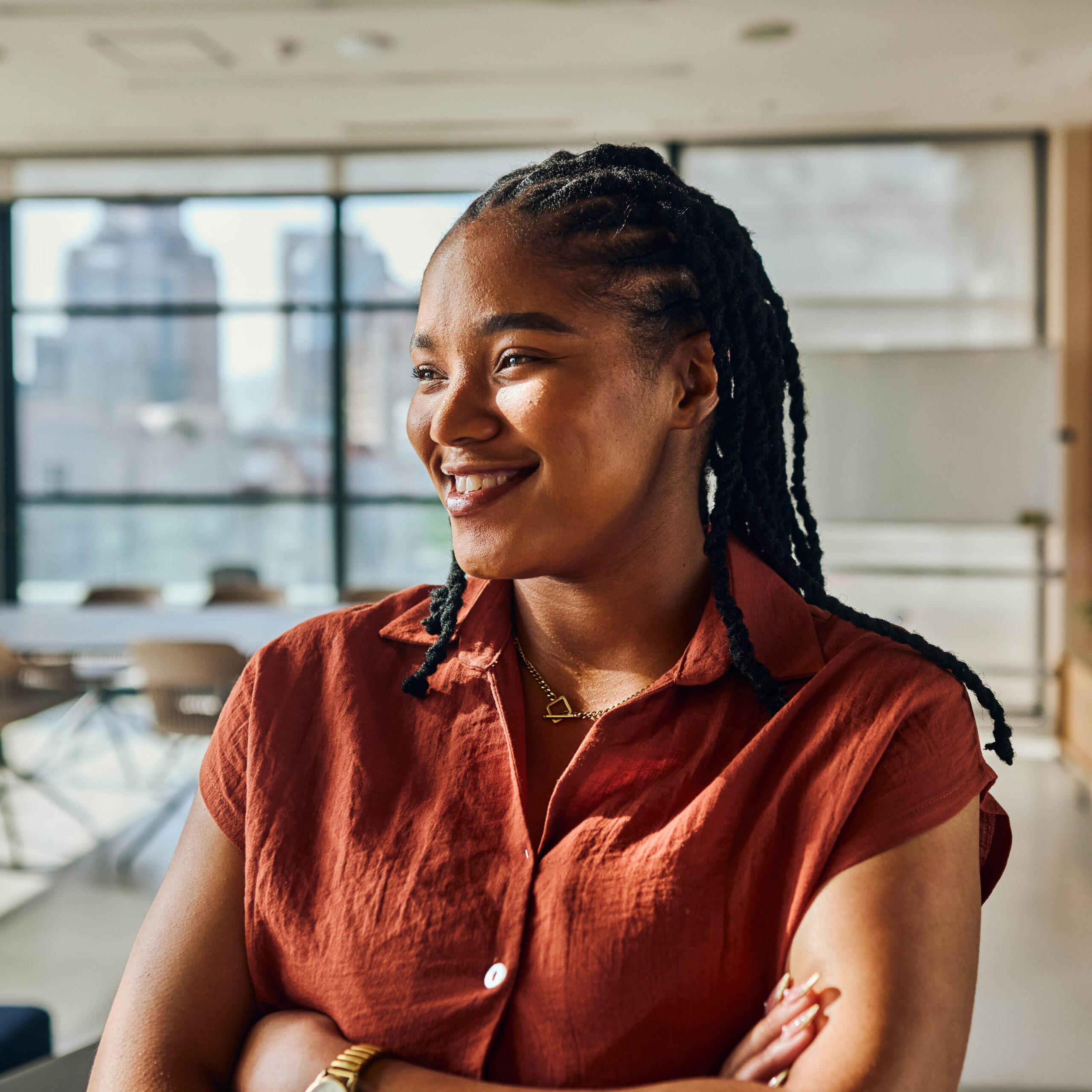 A woman with braided hair wearing a rust-colored short-sleeve shirt stands smiling with arms crossed in a bright, modern office with large windows in the background.