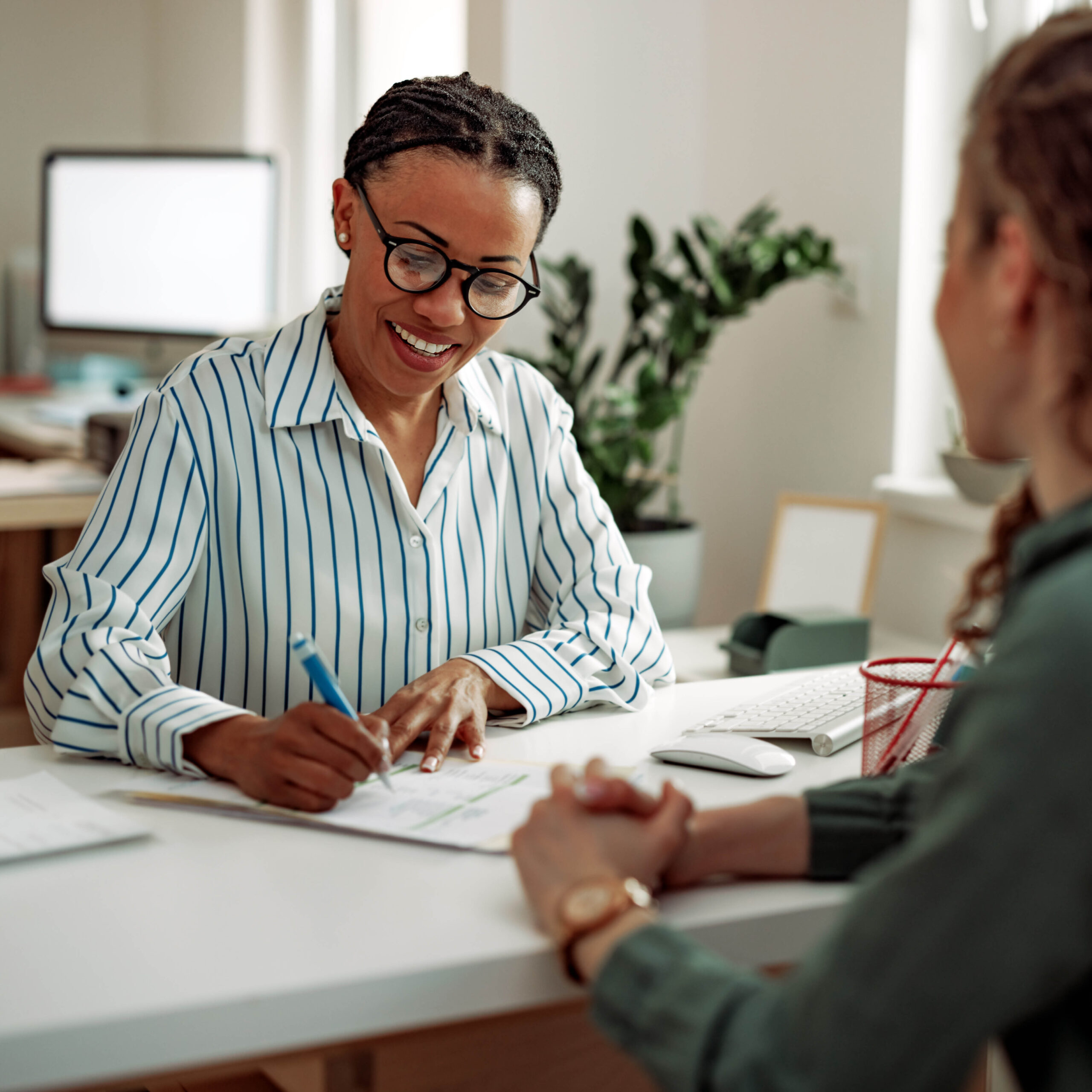 A smiling woman in glasses and a striped shirt writes on a document while talking to another person across a desk in a bright office setting.
