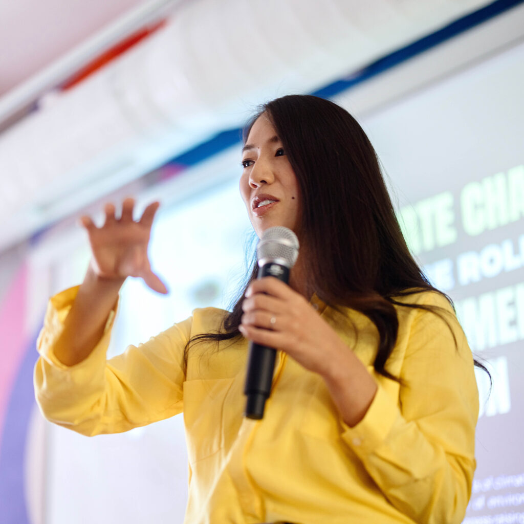 A woman in a yellow shirt speaks into a microphone, gesturing with her hand in front of a projection screen during a presentation.