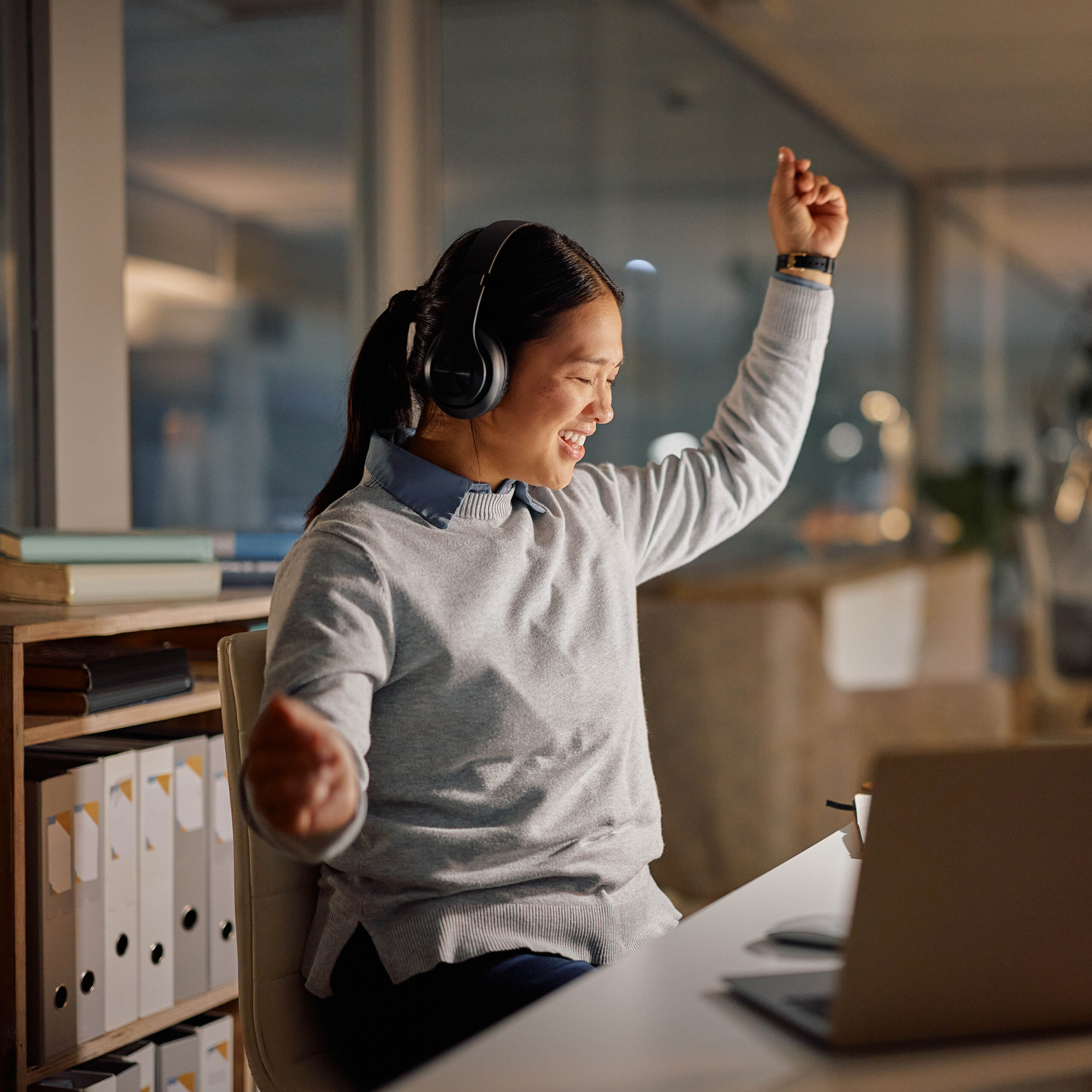 A woman wearing headphones sits at a desk, smiling and raising her arms in excitement while looking at a laptop in an office setting.