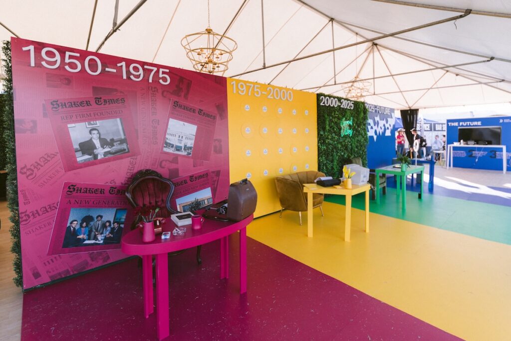 A colorful exhibit displays four time periods—1950–1975, 1975–2000, 2000–2025—each with themed decor and furniture under a white tent, showcasing historical objects and photos on the walls and tables.