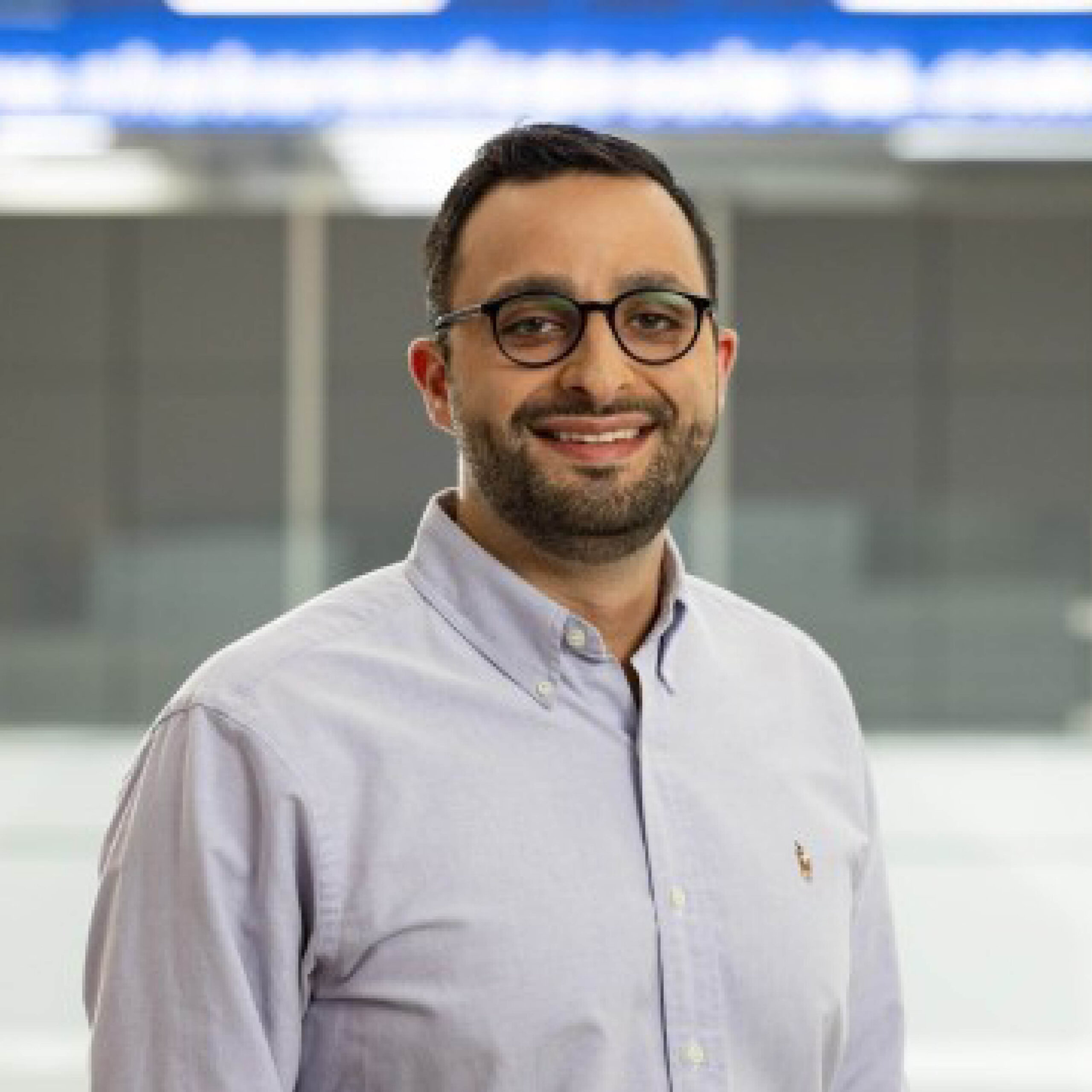 A man with short dark hair, a beard, and glasses is smiling. He is wearing a light blue button-down shirt and standing in a modern, bright indoor setting with blurred background.
