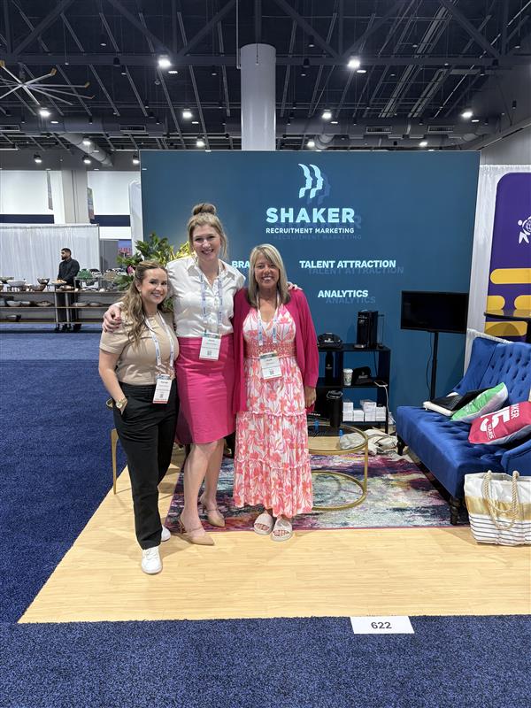 Three women stand and smile in front of a blue Shaker Recruitment Marketing booth at a conference. The booth features a sofa, rug, and marketing materials. Conference attendees and booths are visible in the background.