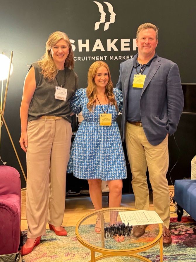 Three people stand and smile together at a conference booth. Two women and one man wear business attire with name badges. The booth backdrop says “SHAKER Recruitment Marketing.” A round table with pens is in front.