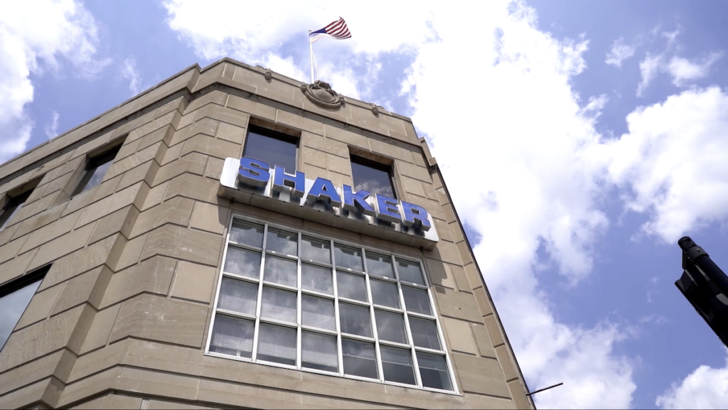 A tan stone building with large windows and the word “SHAKER” in blue letters. An American flag waves on the roof against a partly cloudy sky.