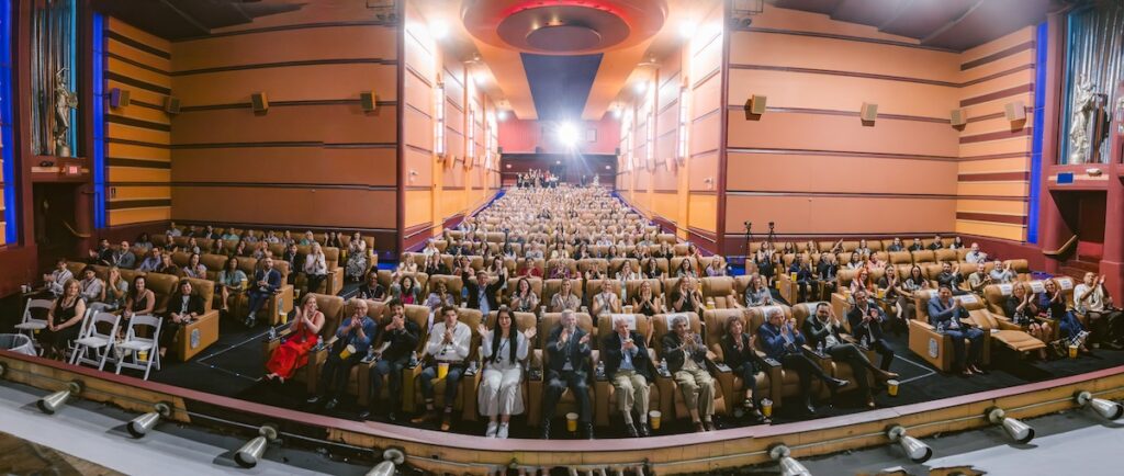 A large audience sits in a theater with beige seats, clapping and facing the stage. The theater features brown and orange walls, bright lights, and a high ceiling.