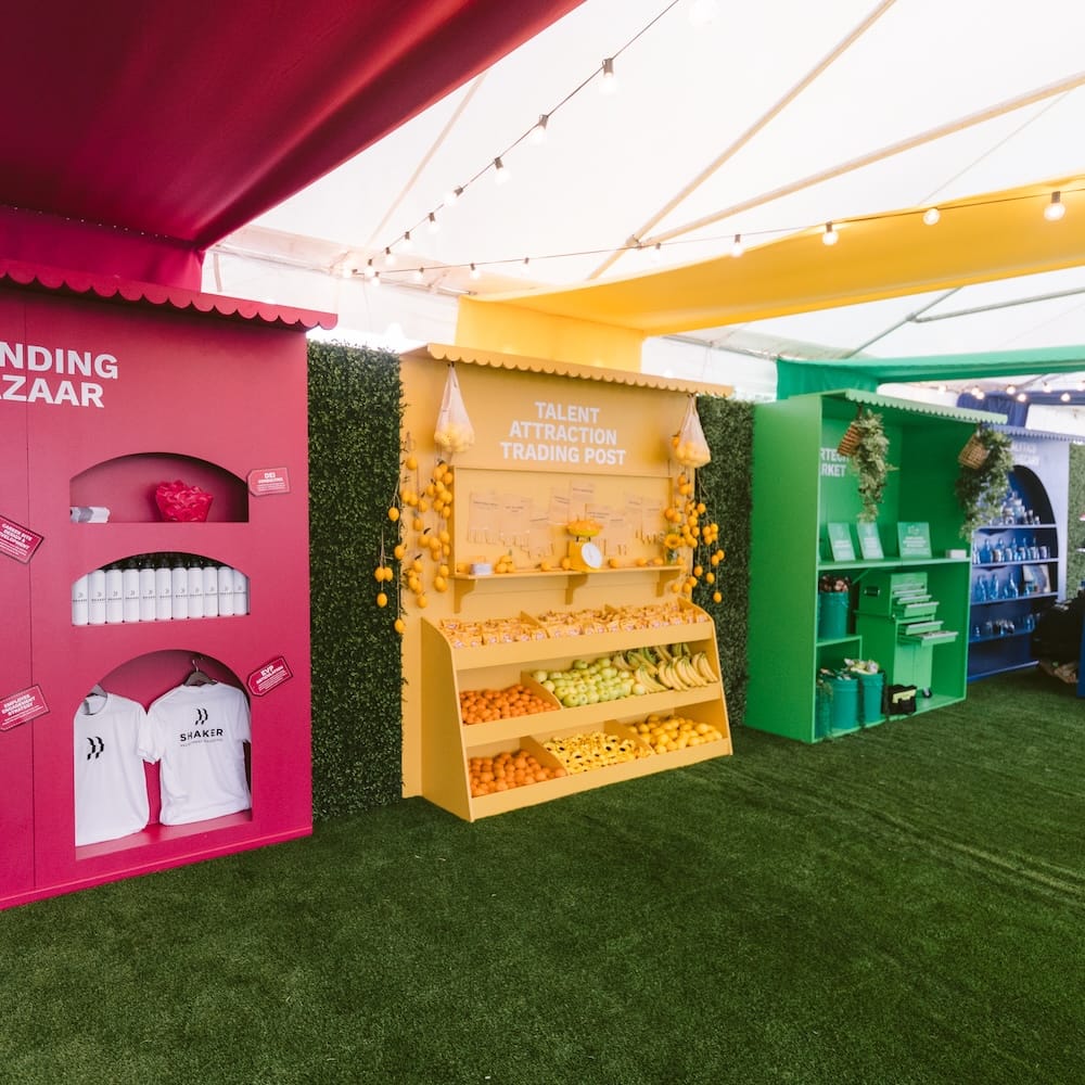 Three colorful booths under a tent display goods including branded t-shirts, produce, and bottles, with a Talent Attraction Trading Post in the center.