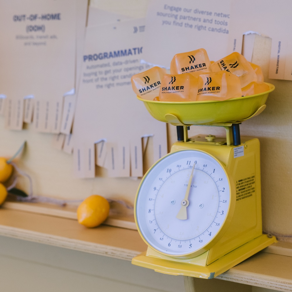 A yellow kitchen scale with orange SHAKER branded items on top, set on a counter with lemons and papers pinned to a bulletin board in the background.
