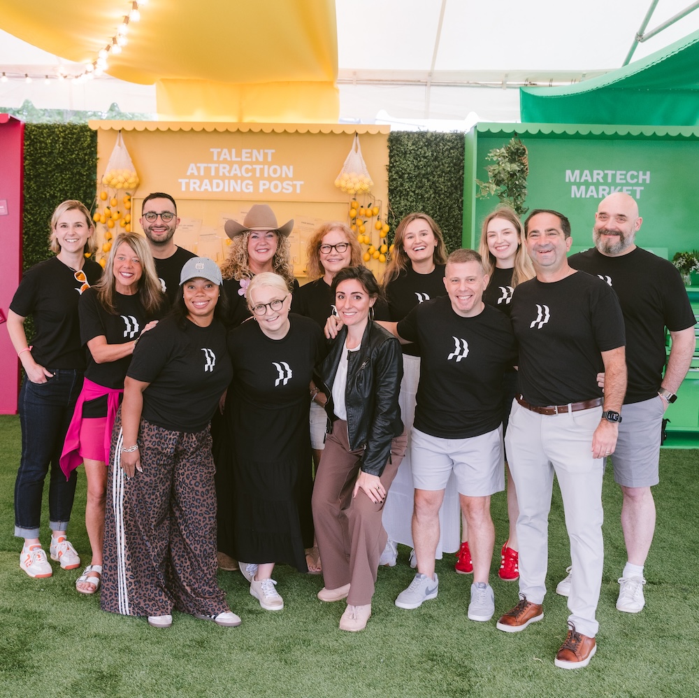 A group of thirteen people, most wearing matching black shirts with white designs, pose and smile together in front of colorful booths at an indoor event.