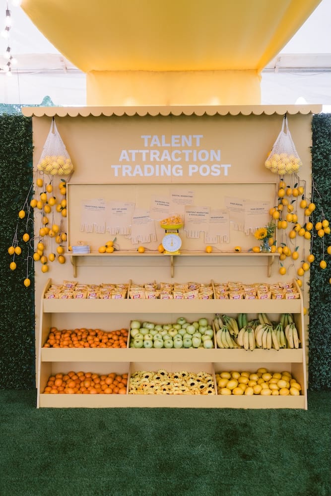 A display titled “Talent Attraction Trading Post” featuring shelves stocked with oranges, green apples, bananas, sunflowers, and packaged snacks, decorated with hanging clusters of lemons.
