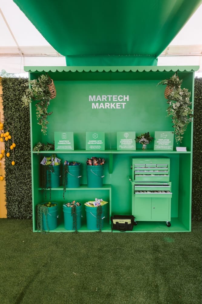 A green “Martech Market” booth featuring four labeled service categories, shelves filled with snack buckets, and a green rolling tool chest.