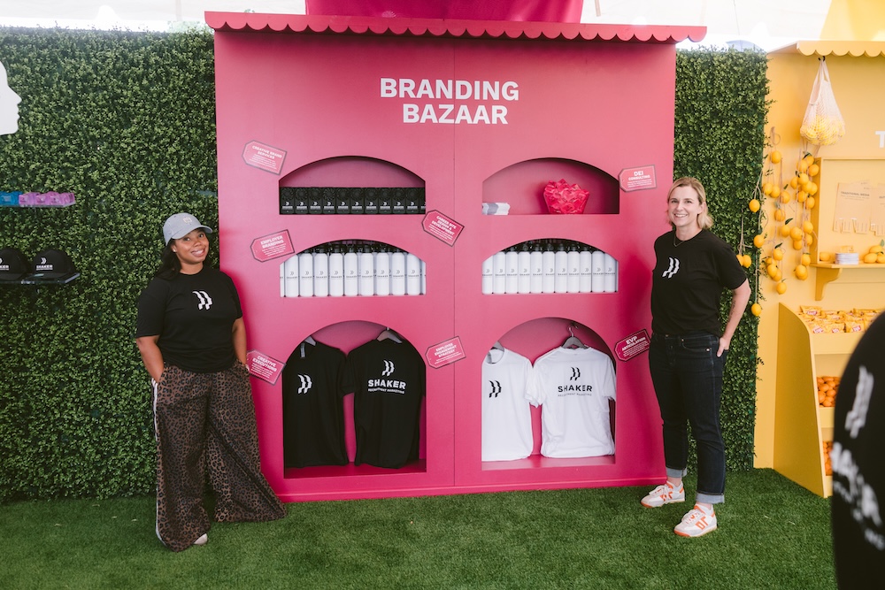 Two women in branded black t-shirts stand in front of a pink “Branding Bazaar” kiosk displaying merchandise like water bottles and shirts, set against a green hedge wall.